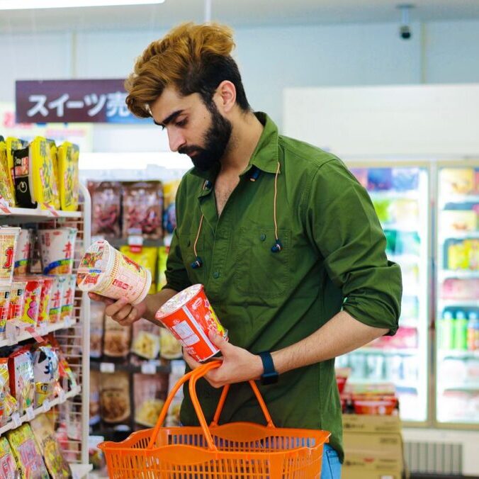 Adult man choosing cup noodles in a vibrant supermarket aisle holding an orange basket.